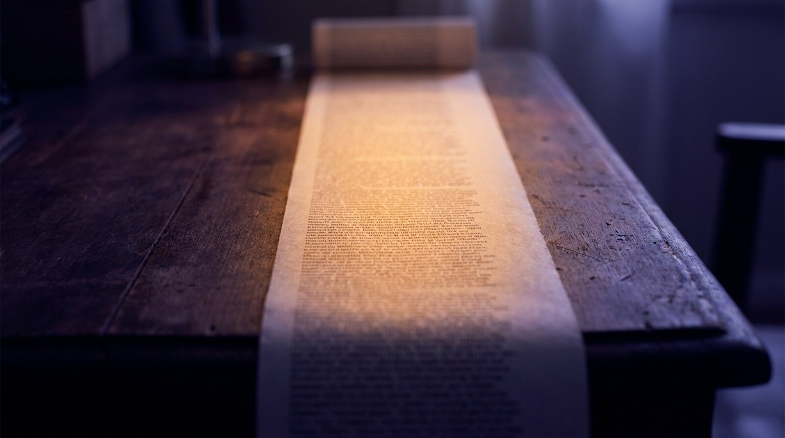A long strip of densely printed paper unrolled flat across a desk, legible only where a band of lamplight falls, both ends lost to shadow